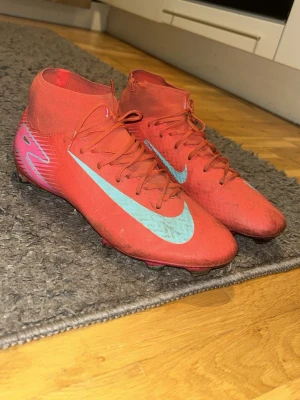Röd/rosa Nike Mercurial fotbollsskor - Säljer ett par röda Nike Mercurial fotbollsskor med högt skaft och blå Nike-logga på sidan. Skorna har snörning och är tillverkade i ett lätt syntetmaterial för bra bollkontroll och grepp på planen. Perfekta för dig som vill ha snabbhet och stil på fotbollsplanen.