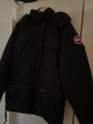 Svart parkas från Canada Goose - Säljer en svart parkas från Canada Goose med klassisk patch på ärmen, fyra stora fickor framtill och hög krage med huva. Jackan har en rak passform och är perfekt för kalla dagar. Materialet är slitstarkt och designen är tidlös.