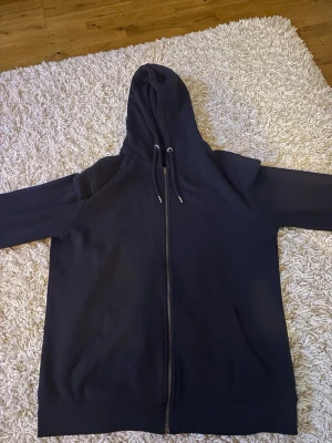 Svart zip hoodie med huva - En enkel och stilren svart hoodie med dragkedja framtill och justerbar huva med snören. Hoodien har långa ärmar och ribbade muddar vid ärmslut och nederkant. Perfekt för en avslappnad och casual look.