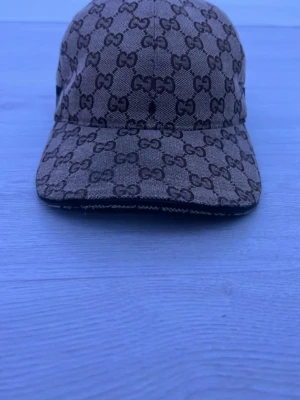 Gucci keps beige - Säljer en beige keps från Gucci med det klassiska GG-monogrammet över hela kepsen. Kepsen har böjd skärm och en grön-röd rand på sidan. Tillverkad i canvasmaterial som ger en exklusiv känsla och streetstyle-vibe.