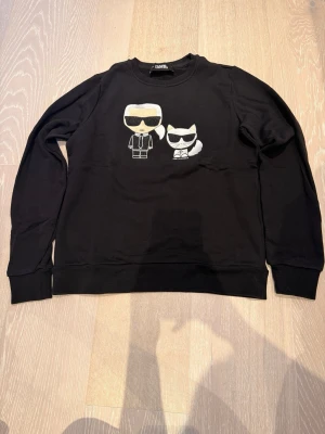 Svart stickad tröja Karl Lagerfeld - Svart tröja från Karl Lagerfeld med coolt broderat motiv av Karl och hans katt på bröstet. Tröjan har rund hals, långa ärmar och ribbade muddar. Perfekt för dig som gillar streetstyle och ikoniska prints.