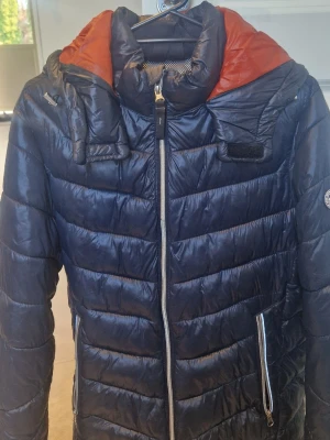 Svart dunjacka från Jack & Jones - Snygg svart dunjacka från Jack & Jones med orange innerfoder och justerbar huva. Jackan har dragkedja framtill, två sidofickor med dragkedja och vattenavvisande material. Perfekt för kalla vinterdagar och riktigt skön att bära.