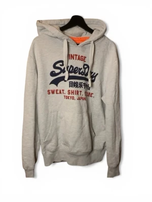 Grå hoodie från Superdry - Ljusgrå hoodie från Superdry med tryck i blått och rött på bröstet samt japanska tecken. Klassisk känguruficka framtill, vita dragsnören i huvan och ribbade muddar. Perfekt för en avslappnad streetwear-look.