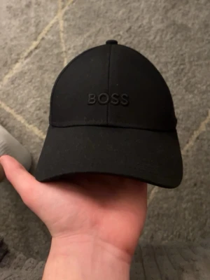Svart keps från BOSS - Snygg svart keps från BOSS med broderad logga framtill. Klassisk böjd skärm och justerbar rem bak för perfekt passform. Tillverkad i ett mjukt och slitstarkt material som känns bekvämt hela dagen. Perfekt accessoar för en clean och stilren look.