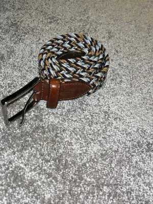 Flätat bälte i blått och brunt - Snyggt flätat bälte med detaljer i brunt skinn och mixade färger i blått, beige och svart. Perfekt accessoar för att lyfta din outfit med en cool och avslappnad vibe. Metallspänne och klassisk design.