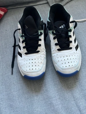 Vita Adidas sneakers med svarta ränder - Snygga vita Adidas sneakers med klassiska svarta ränder på sidan. Skorna har svart plös och svarta skosnören, samt blå detaljer på sulan. Ovandelen är i perforerat syntetmaterial för bra ventilation och komfort. Perfekta för en sportig och avslappnad stil.