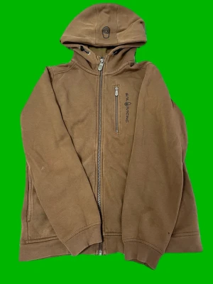 Brun hoodie från Sail Racing - Säljer en brun hoodie från Sail Racing med dragkedja framtill och en bröstficka med logga. Hoodien har huva och ribbade muddar. Perfekt för dig som gillar en stilren och sportig look.