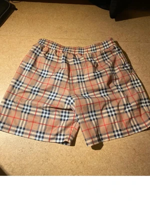 Rutig Burberry shorts i bomull - Säljer ett par klassiska Burberry shorts i beige med det ikoniska rutiga mönstret i blått, rött och vitt. Shortsen har resår i midjan och är tillverkade i mjuk bomull, perfekta för sommardagar.