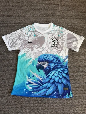 Fotbollströja - Unik Brasilien-tröja från Nike med ett stort blått papegojmotiv framtill. Tröjan har vita och turkosa nyanser, svart Nike-logga och CBF Brasil-märke på bröstet. Tillverkad i ett lätt och ventilerande material, perfekt för sport och träning.