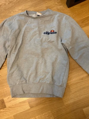 Grå sweatshirt från Ellesse - Klassisk grå sweatshirt från Ellesse med broderad logga i blått, rött och orange på bröstet. Tröjan har rund halsringning, ribbade muddar vid ärmslut och nederkant samt en avslappnad passform. Perfekt för en casual och sportig stil.