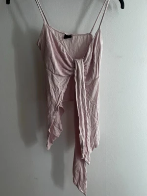 Ljusrosa omlottopp från Gina Tricot - Trendig ljusrosa omlottopp från Gina Tricot med smala axelband och snygg knytning framtill. Toppen har asymmetrisk nederkant och är tillverkad i ett mjukt, tunt material som känns skönt mot huden. Perfekt för dig som vill ha en unik och stilren look. Aldrig använd