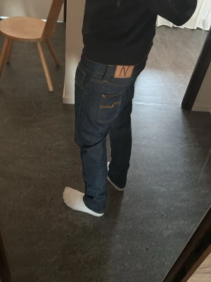 Mörkblå jeans från Nudie Jeans - Säljer ett par mörkblå jeans från Nudie Jeans med klassisk rak passform och kontrastsömmar. Jeansen har tydliga bakfickor med Nudie Jeans signaturbroderi och en läderpatch i midjan. Perfekta för en clean och stilren look.
