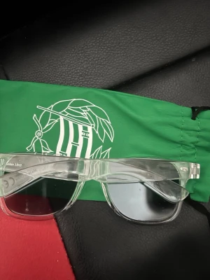 Transparenta solglasögon från hammarby shopen - Säljer ett par stilrena solglasögon med transparent båge och spegelglas. Bågarna har en klassisk form och glasen ger en cool, modern look. Perfekta för dig som vill sticka ut med en unik accessoar. Medföljer grön tygpåse med tryck.