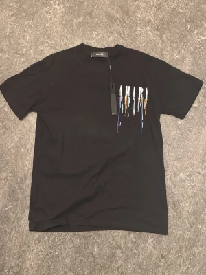 Svart AMIRI t-shirt med färgstänk - Säljer denna svarta t-shirt från AMIRI med korta ärmar och coolt tryck framtill där AMIRI-loggan är dekorerad med färgstänk i blått, orange, grönt och vitt. Tillverkad i mjuk bomull för skön passform. Perfekt för dig som vill sticka ut lite extra. Är storlek S, men passar mer som M.