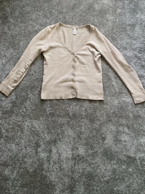 Beige kofta från H&M - Säljer en beige kofta från H&M med v-ringning och knappar framtill. Koftan har långa ärmar och bred ribbad mudd vid ärmslut och nederkant. Perfekt för alla typer av outfits, passar med allt! Finns inte längre att köpa i affär.  Är snygg med linne under men passar även utan!💗
