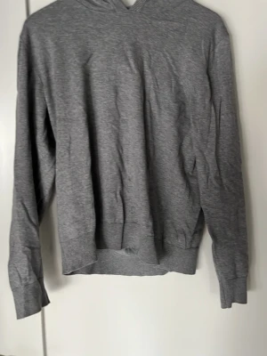 GRUNT Grå hoodie i mjukt bomullstyg - En enkel och stilren grå hoodie med huva och långa ärmar. Tillverkad i mjukt och tunt bomullsliknande material, perfekt för en avslappnad look. Ribbstickade muddar vid ärmslut och nederkant. Passar till det mesta i garderoben.