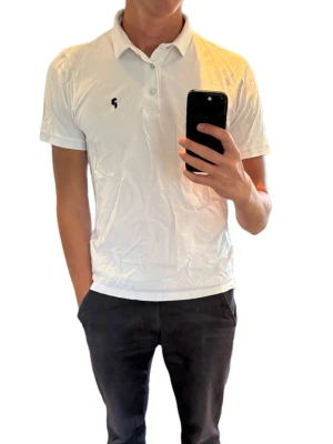 Vit Golfpike från peakperformance med kort ärm - Klassisk vit pikétröja från peakperformance med korta ärmar och krage. Tröjan har knappar framtill och en diskret svart logga på bröstet. Tillverkad i mjuk bomull som känns skön mot huden. Perfekt till jeans eller shorts för en clean look.