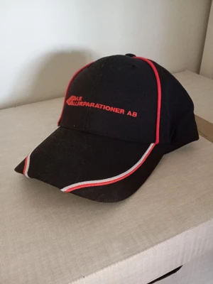Svart keps med röd och vit kant - Snygg svart keps med röda och vita detaljer längs skärmen och panelerna. Framtill finns röd brodyr med texten 'Källe Källreparationer AB'. Klassisk böjd skärm och justerbar passform bak. Perfekt för dig som vill ha en unik och sportig look.