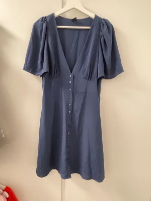 Blå klänning med knappar Gina Tricot - Supersnygg blå klänning från Gina Tricot med v-ringning, korta puffärmar och knappar hela vägen framtill. Klänningen har en flowy passform och är perfekt för sommarens alla tillfällen. Tillverkad i ett mjukt och lätt material.