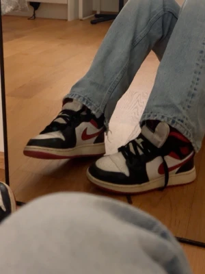 Jordan 1 - Säljer ett par Nike Air Jordan 1 Retro High OG sneakers i svart, vitt och rött. Klassisk high-top modell med ikonisk swoosh på sidan, svart snörning och röd yttersula. Ovandelen är i läder och designen är riktigt tidlös och street.