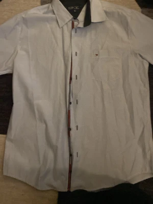 Vit skjorta från Tommy Hilfiger XL - Snygg vit skjorta från Tommy Hilfiger i storlek XL. Skjortan har diskreta tunna ränder, klassisk krage, knappstängning med röd och marinblå detalj samt en bröstficka med liten logga. Perfekt för en clean och stilren look.