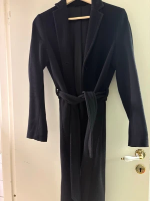 Svart lång kappa med bälte - Stilren svart kappa med klassisk krage och knytbälte i midjan från filippa K. Lång modell med raka ärmar och enkel knäppning framtill. Perfekt för dig som vill ha en tidlös och elegant look.