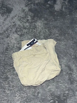 Tommy Hilfiger  Cargo Shorts -  cargoshorts från Tommy Hilfiger i en tidlös beige färg. Shortsen är gjorda i en robust bomullskvalitet och har de karaktäristiska sidofickorna med lock och snygga knappar. De har en avslappnad passform och är perfekta för sommarens alla äventyr. En rejäl och snygg baskomponent i garderoben. • Skick: Mycket fint skick. 