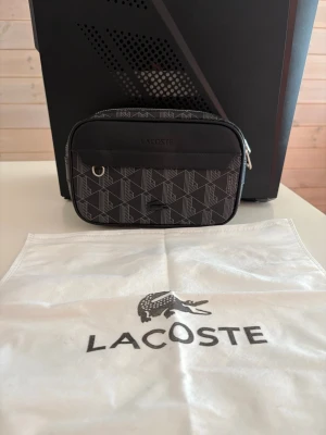 Svart axelväska från Lacoste - Snygg svart axelväska från Lacoste med geometriskt mönster och silverfärgade detaljer. Väskan har justerbar axelrem, dragkedja och en praktisk ytterficka. Tillverkad i slitstarkt material med mesh på baksidan. Perfekt för att bära det viktigaste.