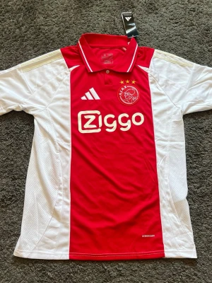 Ajax Amsterdam tröja - Säljer en officiell Ajax Amsterdam-hemmatröja från säsongen 24/25. Tröjan är i storlek Medium (Slim Fit) och är i absolut nyskick med prislappen fortfarande fastsatt. Snygg klassisk modell med krage. 