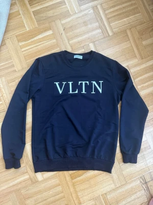 Valentino Tröja - Cool svart sweatshirt med stort vitt VLTN-tryck på bröstet. Tröjan har rund halsringning, långa ärmar och ribbade muddar vid ärmslut och nederkant. Perfekt för en avslappnad och trendig look. Storlek S-M helt ny har aldrig använts.