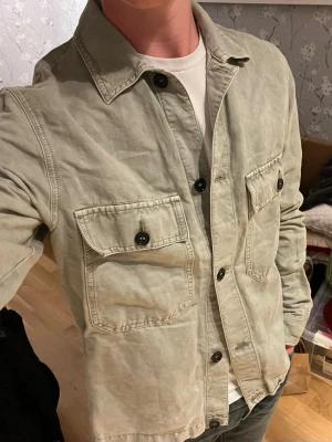 Grön Arket overshirt - Snygg beige overshirt i bomull med två stora bröstfickor med lock och svarta knappar. Rak passform och klassisk krage ger en avslappnad look. Perfekt att bära över en t-shirt för en stilren och enkel outfit.