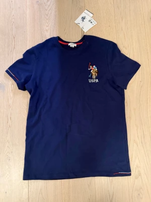 Marinblå USPA t-shirt  - Snygg marinblå t-shirt från USPA med USPA-logga på bröstet. Klassisk rund halsringning och korta ärmar. Tillverkad i mjuk bomull för skön känsla. Diskreta röda detaljer vid ärmslut och nederkant ger extra stil.