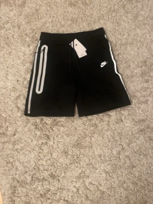 Svarta Nike shorts med reflexdetaljer - Snygga svarta shorts från Nike med vita reflexdetaljer längs sidorna och logga på vänster ben. Shortsen har snörning i midjan och är perfekta för träning eller chill. Tillverkade i mjukt material för maximal komfort.