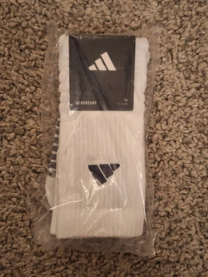 Vita Adidas strumpor AEROREADY - Fräscha vita strumpor från Adidas med svart logga på skaftet. Storlek: 43-45. Modellen är AEROREADY som andas och är perfekta för dig som vill ha bekväma och sportiga strumpor. Ribbad struktur upptill för extra komfort. Skick: 10/10