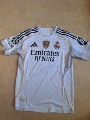 Real Madrid matchtröja Valverde 8 - Säljer en vit Real Madrid matchtröja från Adidas med svarta detaljer och gula linjer. Tröjan har klubbmärke, sponsortryck och Champions League-märke på ärmen. På ryggen står det Valverde och nummer 8. Perfekt för fotbollsfans!