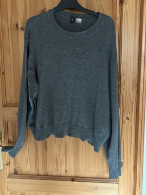 Grå stickad tröja med rund hals - En enkel och stilren grå stickad tröja med rund halsringning och långa ärmar. Tröjan har ribbade muddar vid ärmslut och nederkant, vilket ger en avslappnad och bekväm passform. Perfekt att matcha med jeans eller kjol.
