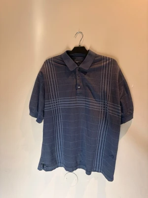 Blå rutig piké från Burberry Golf - Snygg blå pikétröja från Burberry Golf med rutigt mönster och klassisk krage. Tröjan har korta ärmar och knappar framtill. Perfekt för en stilren och sportig look.