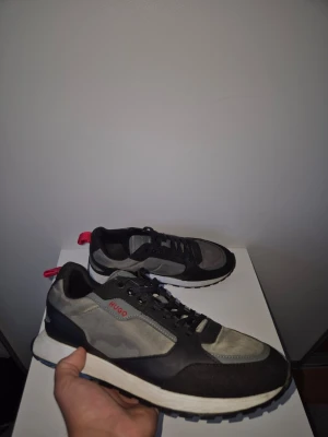 Hugo Skor - Snygga sneakers från Hugo i grått och svart med röda detaljer. Skorna har en klassisk låg siluett, svarta snören och vit sula. Materialmix av mocka och textil ger en modern look. Perfekta för dig som vill ha stilrena och trendiga sneakers.