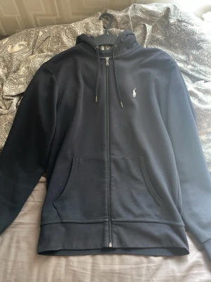 Marinblå zip hoodie från Polo Ralph Lauren - Snygg marinblå hoodie med dragkedja från Polo Ralph Lauren. Klassisk design med huva och dragsnören, två stora fickor framtill och den ikoniska vita logon broderad på bröstet. Perfekt för en avslappnad och stilren look.