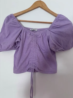 Ljuslila omlottopp med puffärm - Trendig ljuslila topp med puffiga korta ärmar och snörning framtill. Toppen har en omlott-look och croppad passform, perfekt för en modern stil. Tillverkad i ett mjukt tyg som känns bekvämt mot huden. Det passar för xs och s 