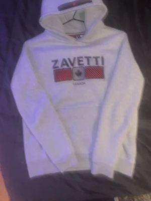 Grå hoodie från Zavetti Canada - Snygg grå hoodie från Zavetti Canada med stor logga och röd-svart detalj framtill. Klassisk känguruficka och huva med dragsko. Perfekt för dig som gillar streetwear och vill ha en stilren look. Mjuk och bekväm bomullsblandning.