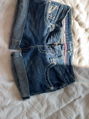 Blå vintage jeansshorts - Säljer ett par vintage blå jeansshorts med vikta ben och kontrastsömmar. Shortsen har flera fickor med dekorativa knappar och detaljer, samt bälteshällor. Perfekta för varma dagar och en avslappnad stil.