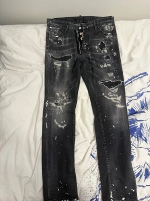 Svarta distressed jeans från Dsquared2 - Snygga svarta jeans från Dsquared2 med slitna detaljer, hål och vita färgstänk. Raka ben och klassisk femficksmodell. Tuff streetstil och tydlig Dsquared2-lapp bak i midjan. Perfekta för dig som gillar en edgy look.