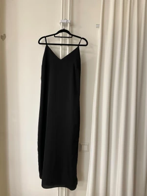 Svart långklänning från VILA - Stilren svart långklänning från VILA med smala axelband och v-ringad hals. Klänningen har en rak och enkel siluett som ger en elegant look. Perfekt för dig som gillar minimalistisk stil.