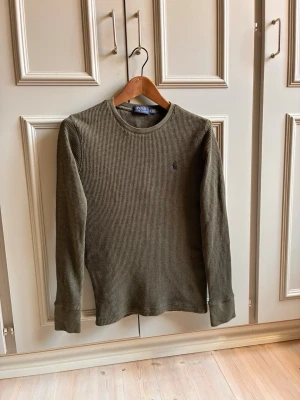 Olivgrön stickad tröja Polo Ralph Lauren - Snygg olivgrön stickad tröja från Polo Ralph Lauren i storlek XS. Tröjan har lång ärm, rund halsringning och ett diskret broderat logomärke på bröstet. Materialet är ribbstickat och ger en skön och avslappnad look.