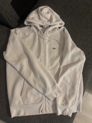 Vit zip-hoodie från Lacoste - Säljer en clean vit hoodie från Lacoste med dragkedja framtill och klassisk krokodil-logga på bröstet. Hoodien har huva med dragsko, två fickor och ribbade muddar. Perfekt för en chill och stilren look.
