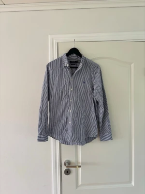 Randig blåvit skjorta från Polo Ralph Lauren - Klassisk långärmad skjorta från Polo Ralph Lauren i blått och vitt randigt mönster. Skjortan har knappar framtill, krage och en liten broderad logga på bröstet. Tillverkad i bomull för en skön och luftig känsla. Skjortan är i storleken 18-20 år i barnstorlek och är självklart äkta. BARA ATT HLRA AV DIG VID FUNDERINGAR! 