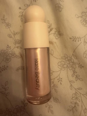 Rare Beauty Enchant Liquid Highlighter - Flytande highlighter från Rare Beauty i nyansen Enchant. Kommer i en glasflaska med rundad vit kork och innehåller 15 ml. Ger ett pärlemorskimrande, ljusrosa glow som smälter in i huden för en fräsch finish.