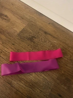 Två träningsband i rosa och lila - Säljer två elastiska träningsband i färgerna rosa och lila. Perfekta för hemmaträning, stretching eller styrkeövningar. Banden är tillverkade i slitstarkt gummimaterial och är smidiga att ta med överallt.