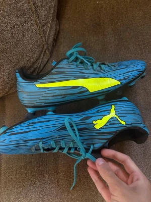 Puma fotbollsskor - Säljer ett par blåa fotbollsskor från Puma med neonfärgade detaljer och logga på sidan. Skorna har snörning, låg profil och mönstrad ovandel i syntetmaterial. Perfekta för dig som vill sticka ut på planen med stilrena och sportiga skor.